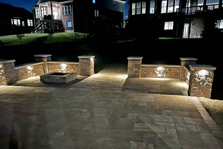 7b9432c5- Illuminated stone patio with low seating walls and a square fire pit at night